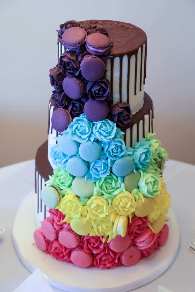 wedding cake image with three tiers and brightly coloured macarons and icing flowers with chocolate drizzle