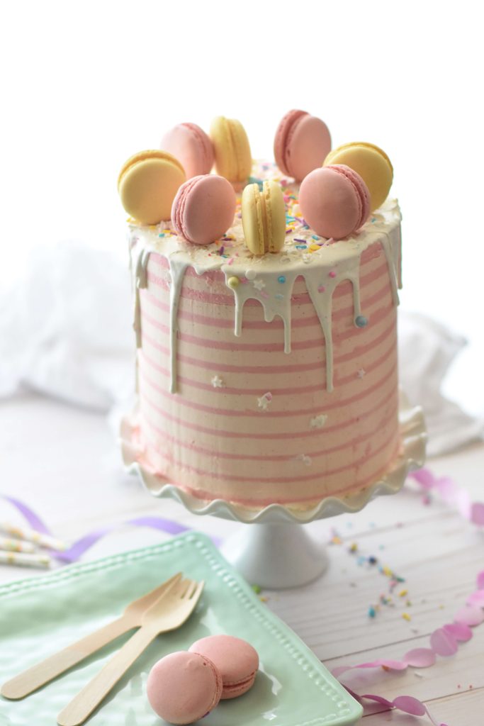 Birthday cake with pastel coloured stripes and macarons on top with white choclate drizzle